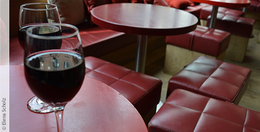 Zwei volle Gläser Wein stehen auf einem rot schimmernden Tisch, im Hintergrund sind ebenso rote Sessel zu erkennen